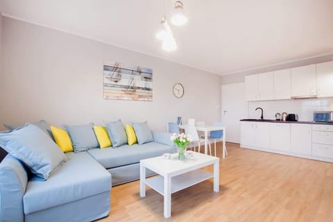 A bright and modern living area featuring a large sofa with colorful cushions, a coffee table, and a wall-mounted TV. Large windows allow plenty of natural light, creating a fresh and welcoming atmosphere.