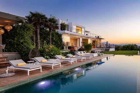 Surrounded by mature trees and fragrant blooms, the setting offers a sense of tranquility and immersion in nature, with sweeping views of the olive groves below and the sea on the horizon