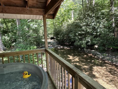 Covered wooden deck with hot tub overlooking a shallow creek surrounded by dense green foliage.
