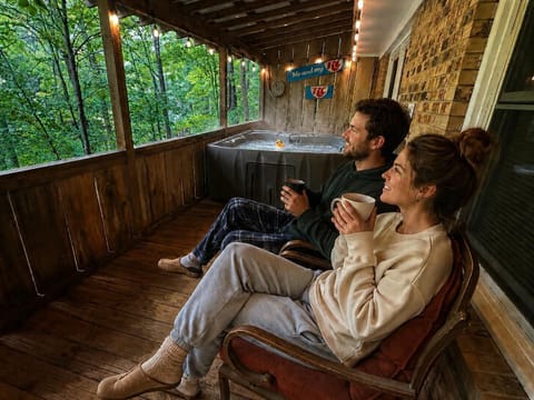 Screened wooden porch with string lights, cushioned vintage chairs, and a hot tub framed by lush green trees.