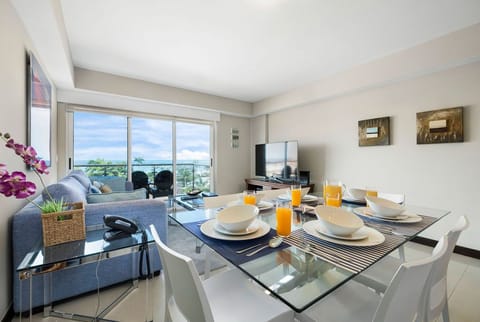 Open-concept living with dining for four, modern entertainment setup, and your private balcony overlooking lush tropical views and the shimmering ocean beyond.