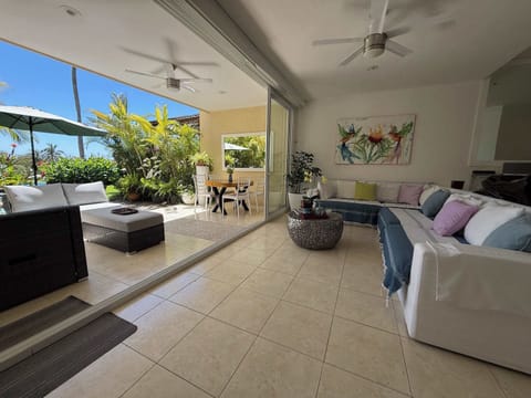 Covered terrace featuring a comfortable lounge area and outdoor dining space, surrounded by lush tropical gardens with partial ocean and pool views.