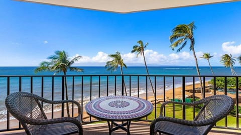 Balcony with mosaic table and chairs overlooking a sandy beach and ocean, palm trees swaying under a clear sky with a few clouds.