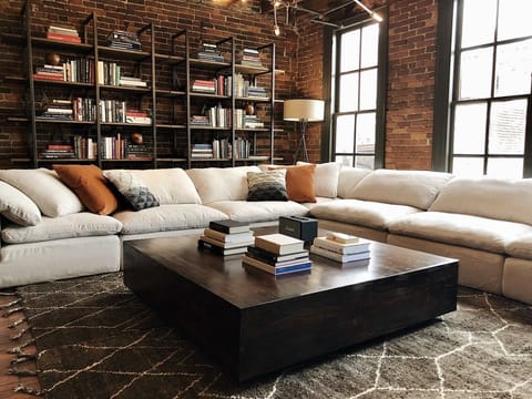 - Plush sectional paired with warm accents for a calm lounge   - Bookshelf wall and tall windows adding charm and texture   - Cozy corner perfect for unwinding in an inviting loft
