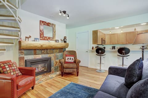 Living room with wood flooring, a stone fireplace with a log mantel, a brown armchair, a red armchair, a navy sofa, a blue rug, and an open kitchen with bar stools. Spiral staircase in the corner.