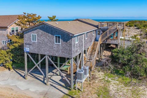 Coastal beach house elevated on stilts with weathered shingle siding, surrounded by natural dune vegetation and ocean views beyond.