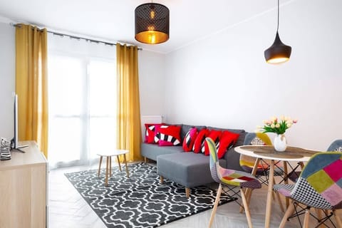 A modern, bright living room with a cozy setup. There are yellow curtains, a small coffee table, and colorful cushions. The room has large windows that let in a lot of natural light.