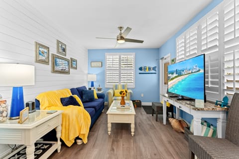 Bright coastal living space with plantation shutters and a wall-mounted smart TV, opening seamlessly into a fully equipped white kitchen — perfect for morning coffee before hitting the sand.