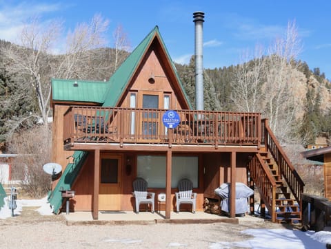 A charming A-frame cabin nestled in a mountain valley, featuring a distinctive green metal roof and wrap-around deck. The property sits surrounded by trees and hillsides, offering a peaceful retreat in a natural setting.