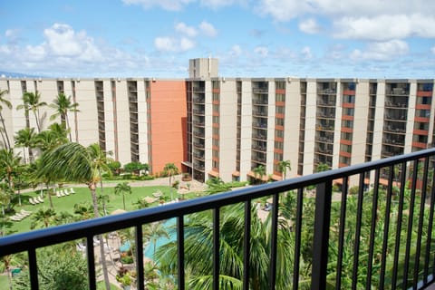 The property features a multi-story building with balconies overlooking a central courtyard with tropical landscaping, a swimming pool, and relaxation areas, all under clear blue skies.