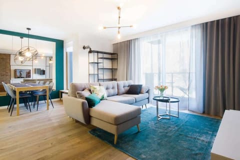 A stylish living room with a modern aesthetic, featuring a cozy beige sofa, vibrant teal rug and coffee table. The room is well-lit with large windows and decorated with tasteful wall art.
