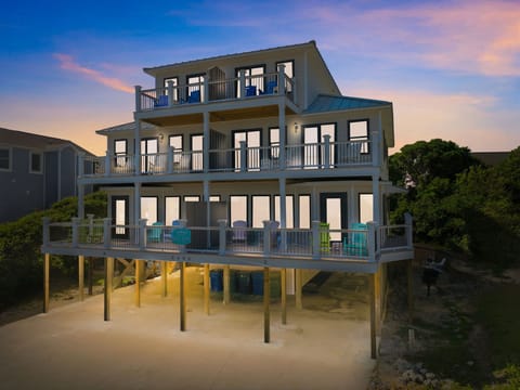 A stunning beachfront retreat elevated on stilts captures a spectacular sunset backdrop, featuring three levels of spacious balconies and panoramic windows overlooking the coastal landscape and surrounding area.