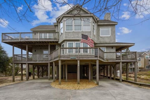 A distinctive coastal property elevated on sturdy pilings, featuring wraparound decks and classic shingle siding. The American flag marks the welcoming entrance to this beachside retreat, set among native coastal trees and just steps from other vacation ho
