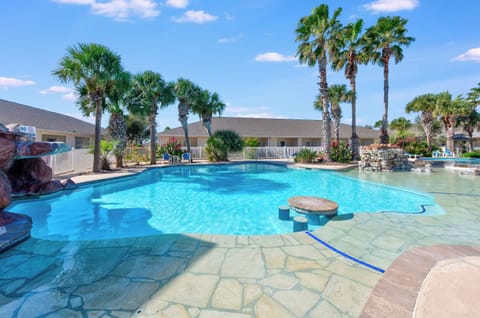 Tropical resort pool area featuring crystal-clear water, palm trees, and comfortable seating islands surrounded by low-rise buildings.