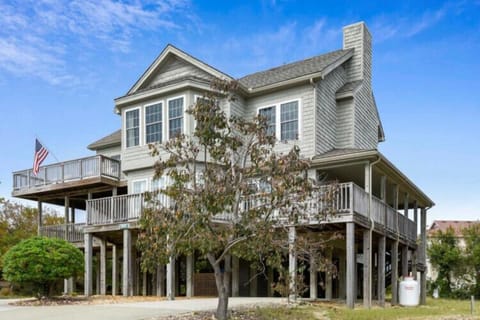 A stately coastal vacation home with multi-level decks and an American flag, surrounded by natural landscaping and positioned for easy beach access.