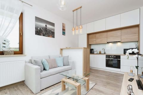 A bright, modern living room with a light-colored sofa, small coffee table, and open-plan design connected to a kitchen. Soft colors and minimalistic decor add a cozy touch.