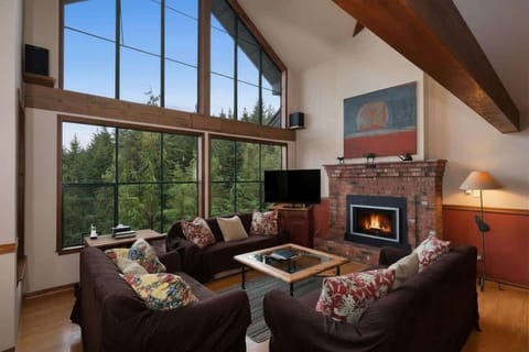 - Cozy living room with high windows framing forest views and a warm brick fireplace  
- Plush seating and exposed beams create the ideal mountain gathering space  
- Relax with movies, cocoa, or games after exploring Whistler’s trails  
