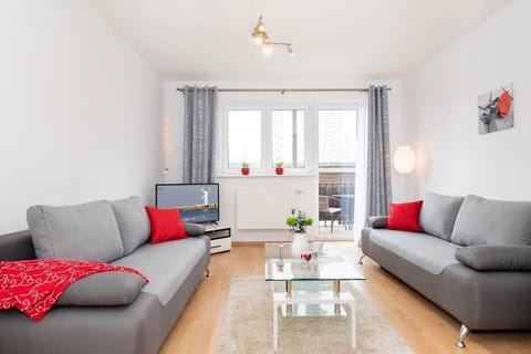 A bright living room with a gray sofa and a red accent pillow, complemented by a coffee table and wooden flooring. The room has large windows letting in plenty of natural light.