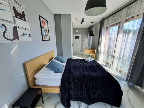 A cozy bedroom featuring a neatly made bed with dark bedding, a desk with a chair, and a large window letting in natural light.