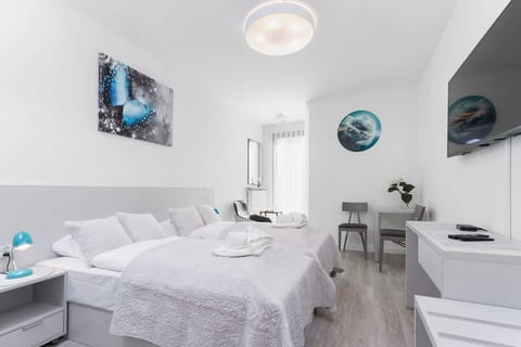 A bright bedroom with a double bed, two bedside tables, and a wall-mounted TV. The decor features modern art and white bedding.