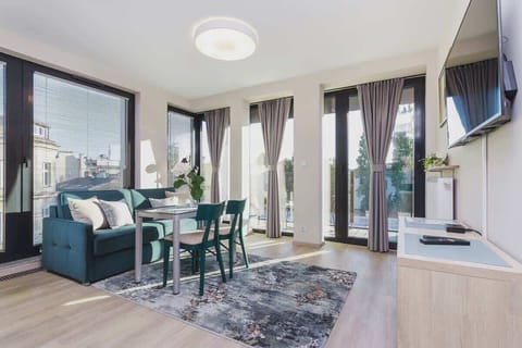 A bright, modern living room with large windows. The space features a dining table with chairs and a cozy seating area with a sofa, showcasing natural light and city views.