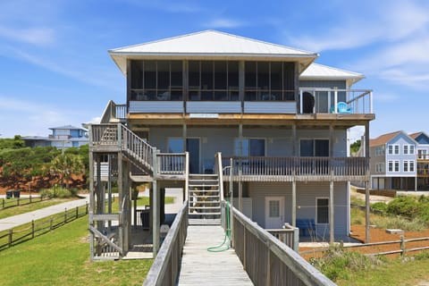 Elevated coastal beach house with multiple levels, screened porch, expansive deck spaces, and weathered wood boardwalk leading to the entry—ideal for a seaside vacation retreat.
