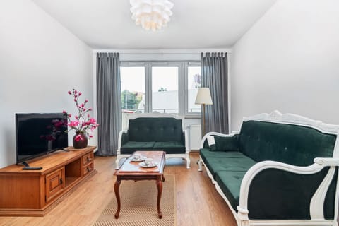 A spacious living room featuring a green upholstered sofa, a wooden coffee table, and a TV cabinet. Large windows with curtains allow natural light to brighten the space.