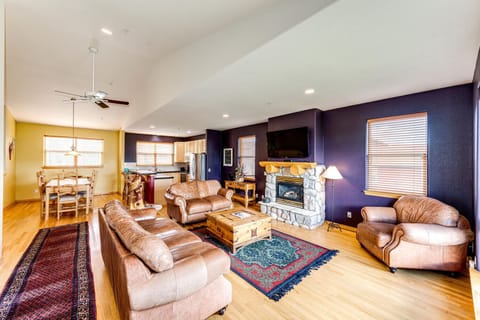 A spacious living room with wooden flooring, featuring leather sofas, a stone fireplace, a wooden coffee table, and a wall-mounted TV. There's a dining area and kitchen visible in the background.