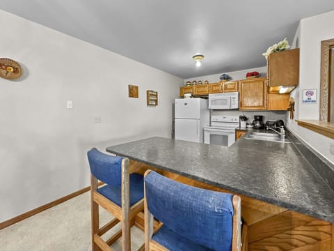 A kitchen with wooden cabinets, a white refrigerator, microwave, and stove. A black countertop has two blue cushioned barstools. A "Home Sweet Home" sign is on the wall.