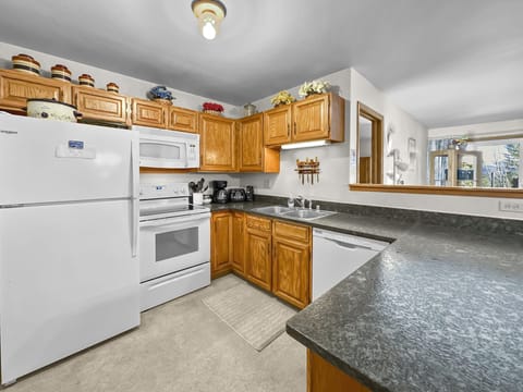A kitchen with wooden cabinets, a white refrigerator, stove, microwave, and a dishwasher. Countertops are dark granite with appliances like a toaster and coffee maker. There is a pass-through window.