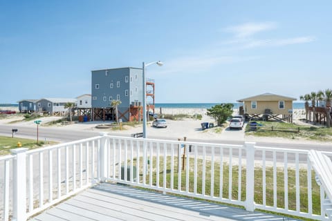 View from a beach house deck overlooking direct beach access in Oak Island, NC. A sandy path leads across the dunes to the ocean, with neighboring coastal homes, parked cars, and bright blue skies framing the shoreline.