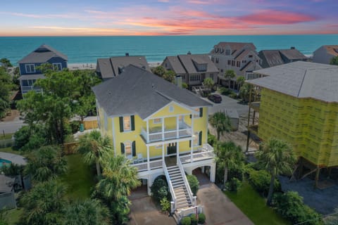 Charming yellow beach house nestled among coastal homes with stunning ocean views and tropical palm landscaping.