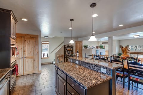 A modern kitchen with dark wood cabinetry, granite countertops, stainless steel appliances, and a double sink. A clock hangs on the wall above a window. A pendant light fixture is visible.