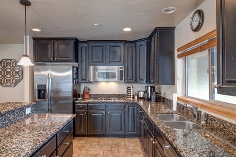 A modern kitchen with dark wood cabinetry, granite countertops, stainless steel appliances, and a double sink. A clock hangs on the wall above a window. A pendant light fixture is visible.