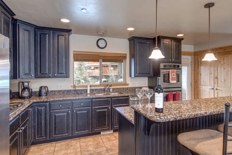 A modern kitchen with dark wood cabinetry, granite countertops, stainless steel appliances, and a double sink. A clock hangs on the wall above a window. A pendant light fixture is visible.