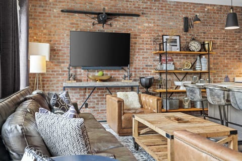 Stylish open-concept living room featuring exposed brick walls, comfortable leather seating, a large Smart TV, rustic wood accents, and a seamless flow into the fully equipped kitchen and dining area.
