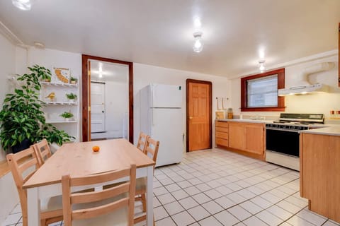 A spacious eat-in kitchen with tiled floors, wood cabinetry, full-size appliances, dining table, greenery, and bright overhead lighting.
