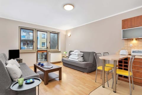 The spacious living room features a grey sofa set and a coffee table, with warm wooden flooring. Large windows allow ample natural light, creating a cozy and bright atmosphere.