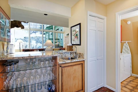 A home bar area with granite countertops features a sink, glass shelving filled with various glassware, and light wood cabinetry, adjacent to a hallway leading to a laundry room with a washer and dryer.