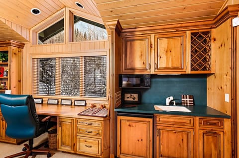 A home office with wooden cabinets, a desk, a blue office chair, a small sink, and a microwave. A window with blinds reveals a snowy outdoor scene.