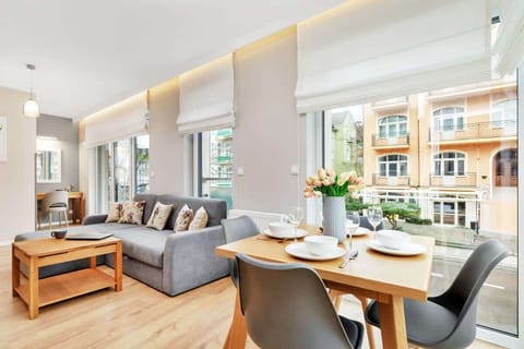 A wider perspective of the living room, including the sofa, a wooden dining table, and bright natural light streaming in through the windows.