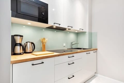 The compact kitchen features sleek white cabinets, a glass backsplash, and essential appliances like a microwave and kettle. The clean and modern design prioritizes functionality.