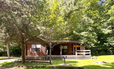 Front view of the log cabin surrounded by mature trees, featuring a covered porch with wooden railing and a charcoal grill.