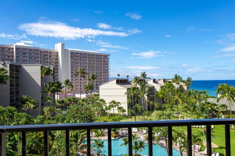 An inviting view from your private balcony reveals a sparkling pool, lush tropical gardens swaying with palm trees, and the stunning Pacific Ocean shimmering on the horizon under brilliant Hawaiian sunshine.