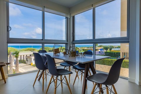 A modern dining space frames turquoise waters and coastal greenery through floor-to-ceiling windows, where your morning coffee meets the perfect view.