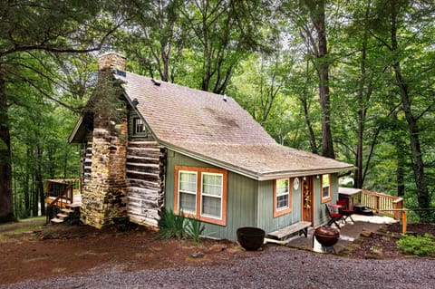 Exterior view of the cabin showing a stone chimney, green wood siding, gravel driveway, and wooded surroundings.