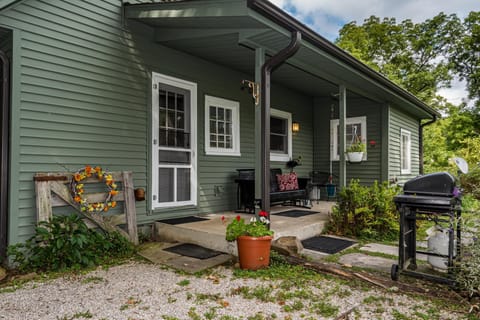 Green siding and white-trimmed windows frame the covered porch with black bench seating and a hanging flower pot.