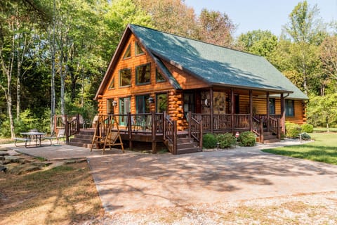 Log cabin exterior with green shingled roof, wraparound porch, and stone fire pit patio surrounded by forest.