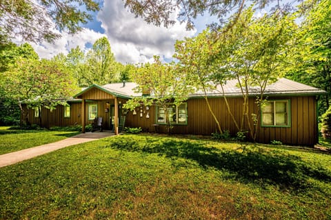 Exterior view of brown wood-paneled house with green window trim, a covered porch, and surrounding leafy trees.