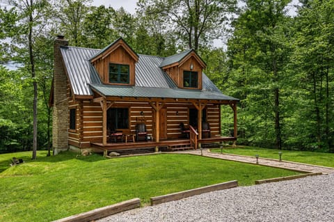 Exterior view of a log cabin with a green metal roof, stone chimney, wooden porch, and rocking chairs.